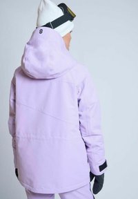 Lavendel skijakke med hætte, der har et geometrisk synsdesign og sorte velcro-detaljer. Båret med handsker og en hvid beanie.