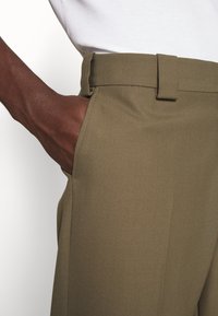 Olijfgroene broeken van gestructureerde stof, met een platte voorkant, ceintuurlussen en zijzakken, met een close-up van de tailleband.