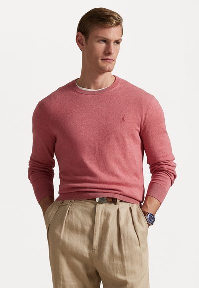 Lichtroze gebreide trui met ronde hals en geribde manchet en zoom, gecombineerd met beige geplooide broek en een zilveren horloge, die een casual pasvorm laat zien.