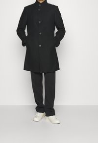 Zwarte, lange woljas met hoge kraag, knoopsluiting aan de voorkant, zakken opzij, gecombineerd met zwarte broek en witte sneakers. Gladde textuur.