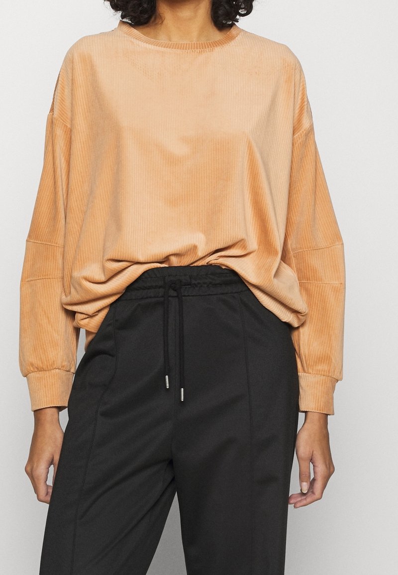 Femme portant un haut beige côtelé à manches longues et ample, rentré dans un pantalon noir taille haute à cordon, sur un fond uni.