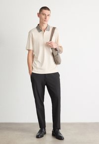 Beige polo skjorte med teksturert krage, kombinert med svarte bukser og svarte sko. Modellen bærer en liten grå skulderveske.