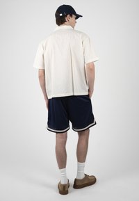 Homme portant une chemise blanche à manches courtes rayée, un short bleu marine, des chaussettes blanches, des chaussures marron et une casquette noire, debout les mains dans les poches.