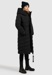 Zwarte pufferjack met een lange, slim fit ontwerp, horizontaal gestikt patroon, oversized capuchon en ritssluiting aan de voorkant. Gemaakt in combinatie met zwarte enkellaarzen.