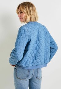 Chaqueta bomber de mezclilla azul acolchada con puños y cintura de canalé, que presenta un patrón texturizado de formas superpuestas. Se muestra vista trasera.
