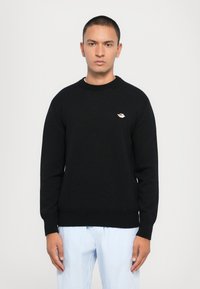 Svart genser laget av teksturert strikket stoff, med rund hals og en liten brodert logo på venstre side. Kombinert med lysstripete bukser.
