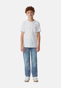 T-shirt en coton blanc avec impression graphique sur le côté gauche, associé à un jean bleu clair et des baskets blanches avec des semelles beige.