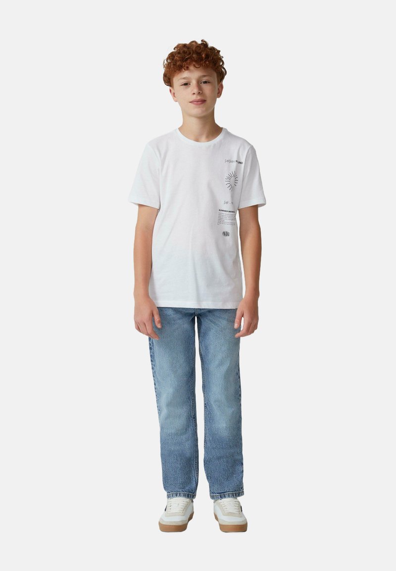T-shirt en coton blanc avec impression graphique sur le côté gauche, associé à un jean bleu clair et des baskets blanches avec des semelles beige.