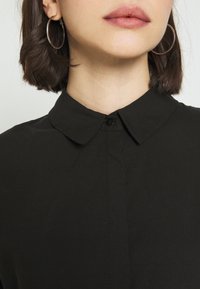 Chemise noire à col avec fermeture à boutons, confectionnée en tissu lisse. Le modèle porte de grandes boucles d'oreilles et a les cheveux courts et foncés visibles.