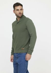 Grüner, strukturierter Pullover mit Schalkragen und gerippten Bündchen. Hat einen kleinen braunen Akzent am Saum. Wird mit blauen Jeans kombiniert.