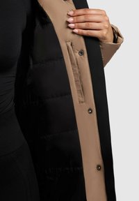 Geaca puffer neagră și bej are o textură exterioară netedă, nasturi tip snap și un buzunar lateral, cu o căptușeală puffer izolată vizibilă pe interior.