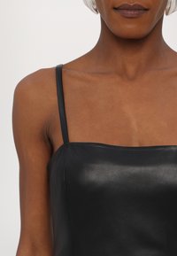 Camisola din piele neagră cu bretele subțiri și decolteu drept, având o textură netedă și un design minimalist. Fără modele sau detalii suplimentare.