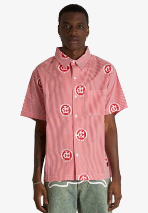 Modello maschile che indossa una camicia a maniche corte con bottoni a righe verticali rosse e bianche, con loghi circolari rossi e jeans azzurri chiari.