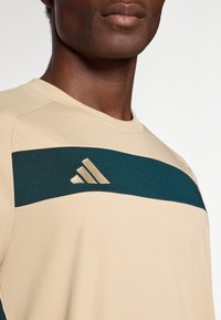 Beżowa koszulka sportowa z poziomą ciemnozieloną paskiem. Posiada złote logo Adidas na pasku. Gładki, teksturowany wzór materiału.