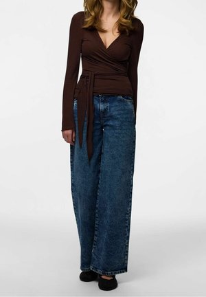 Kvinde iført en mørkebrun slå-om bluse med lange ærmer, bredt bånd og løse, mørkeblå, højtaljede denimjeans samt sorte sko.