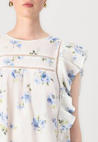 Blusa bianca floreale con fiori azzurri chiari, maniche a rouches delicate e dettagli in pizzo lungo la parte anteriore. Tessuto morbido e leggero.