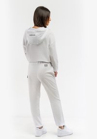 Biela skrátená mikina s kapucňou a šnúrkou, v kombinácii so zodpovedajúcimi slim-fit teplákmi. Má logo "ICE" na vrecku na zadnej strane a príjemnú textúru.
