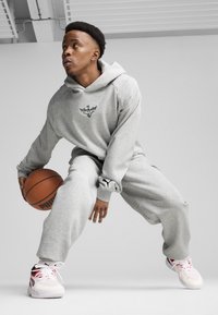 Grå hoodie og joggebukser lavet af blødt stof, med et logo på brystet og et Puma-emblem på venstre ærme. Han holder en basketball.