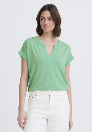 Vrouw die een lichtgroene blouse met korte mouwen en V-hals draagt, ingestopt in een witte high-waisted spijkerbroek, staand tegen een effen achtergrond.
