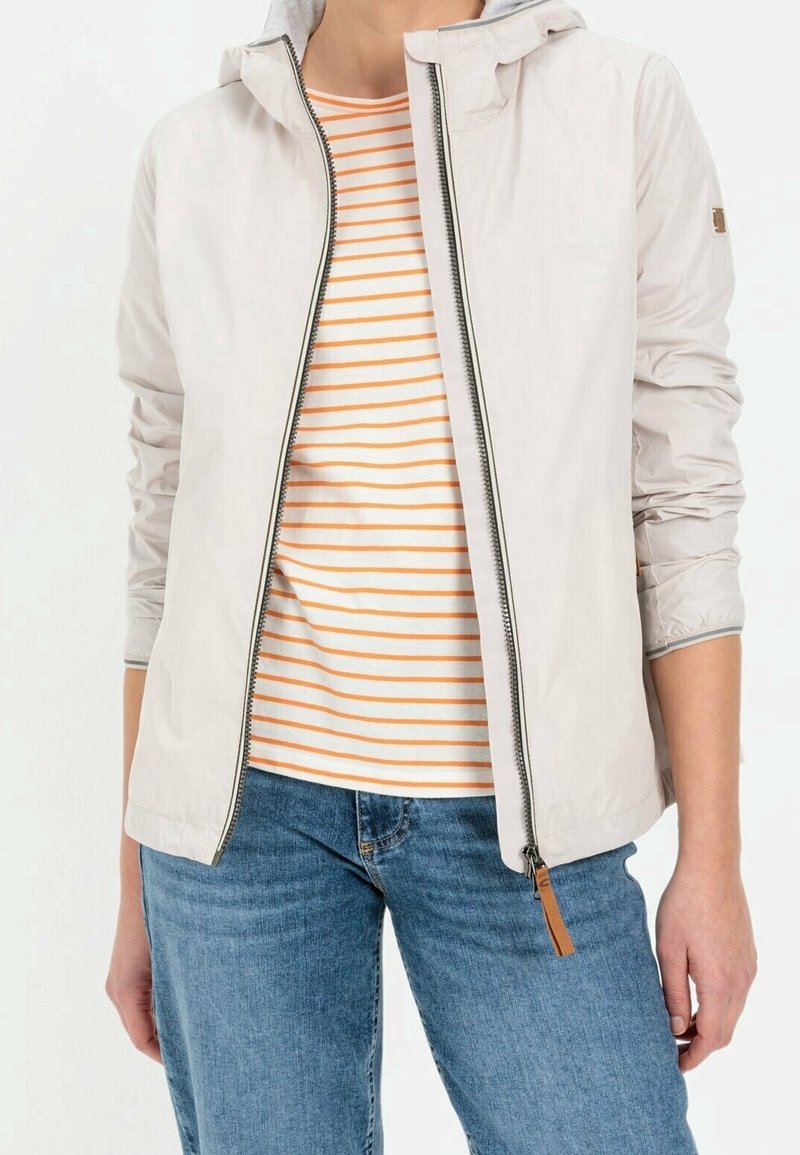 Personne portant une veste à capuche beige clair zippée par-dessus une chemise rayée blanche et orange avec un jean bleu, debout devant un fond blanc.