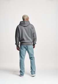 Szara, oversize bluza z kapturem i sznurkiem, zestawiona z jasnoniebieskimi, prostymi jeansami. Widok od tyłu ukazuje miękką fakturę i luźne dopasowanie.