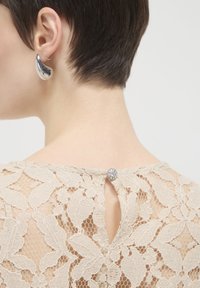 Beige Spitzeoberteil mit floralen Mustern, Schlüsselloch-Rücken und einem dekorativen Knopf. Glatte, zarte Textur mit filigranen Spitze-Details.