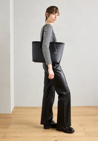 Kvind i sorte læderbukser og stribet langærmet skjorte, der bærer en stor sort mønstret totebag, stående på et lyst trægulv.