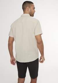 Camicetta in lino a maniche corte di colore beige con orlo dritto e colletto standard, abbinata a pantaloni corti scuri, vista da dietro.
