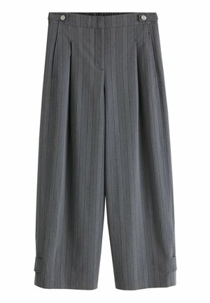Junge Frau mit dunkler Sonnenbrille, grauem Langarmshirt, weitem grauen Hosen und Sandalen, die an einer gefliesten Betonwand steht.