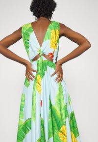 Blomstret ærmeløs kjole med blå bund, store grønne blade og gule bananmønstre, åben ryg med knudedetaljer ved taljen.