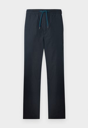 Giovane uomo con capelli ricci che indossa scarpe da ginnastica bianche, pantaloni neri larghi e una maglietta bianca con quattro disegni colorati di teschi.