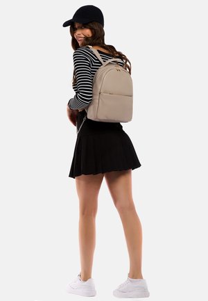 Beige læder rygsæk med kurvet design, guldfarvede hardware og frontlomme. Modellen bærer sort plisseret nederdel og stribet langærmet top.