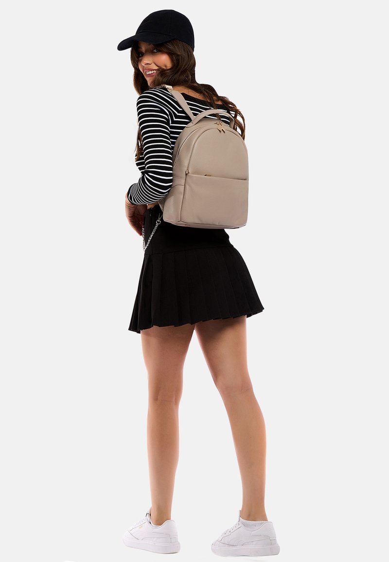 Zaino in pelle beige con design curvo, hardware dorato e tasca frontale. La modella indossa una gonna nera plissettata e una maglietta a righe a maniche lunghe.
