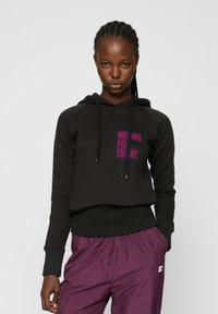 Sudadera negra con capucha y texto rosa "WAITING FOR FRIDAY", con un corte relajado, bolsillo canguro y tejido texturizado. Combinada con pantalones morados.