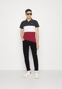 Camisa polo en negro, blanco y burdeos con mangas cortas y cuello, combinada con jeans negros y zapatillas blancas. Los accesorios incluyen gafas de sol.