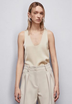 Top in satin beige con scollatura a V profonda e spalline larghe, abbinato a pantaloni plissettati beige chiaro, su uno sfondo neutro.