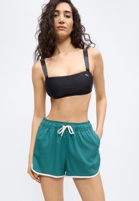 Γυναίκα με μπούκλες που φοράει μαύρο αθλητικό σουτιέν με ράντες Puma και αθλητική βερμούδα τιρκουάζ με κορδόνι στη μέση και λευκό περίγραμμα.