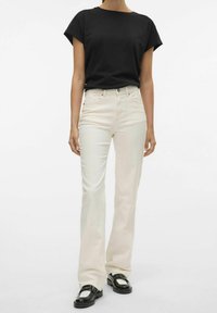 Camiseta negra de manga corta combinada con unos pantalones de mezclilla de tiro alto, de corte acampanado, en color beige claro. La modelo lleva zapatos negros con acentos blancos.