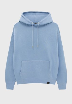 Personne portant un sweat à capuche bleu clair, un jean bleu et des baskets blanches, debout avec les mains dans les poches du sweat à capuche devant un fond uni.