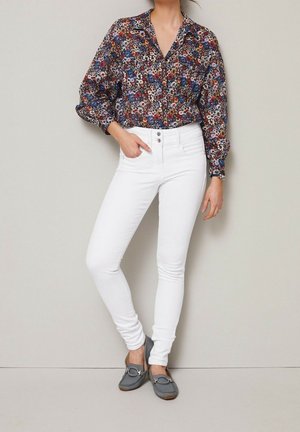 Frau trägt eine langärmlige Blumenbluse, weiße High-Waist Skinny Jeans, graue Loafers, steht vor einer hellgrauen Wand.