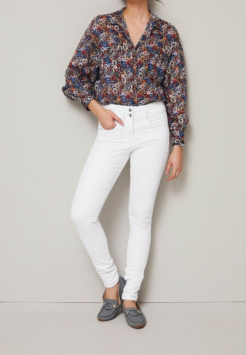 Femme portant un chemisier à fleurs à manches longues, un jean skinny blanc taille haute, des mocassins gris, debout contre un mur gris clair.