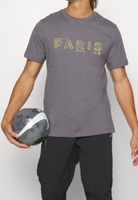 Osoba v šedém tričku s nápisem "PARIS" a černých kalhotách, držící šedobílý fotbal Nike v levé ruce, na jednolitém pozadí.