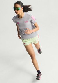Conjunto de running que incluye una camiseta gris con detalles y logo en rosa, pantalones cortos verdes claros con marca y gafas de sol reflectantes.