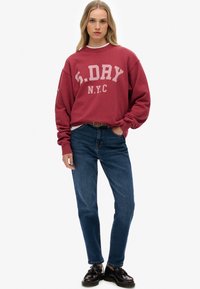 Young woman wearing a red S.DRY NYC sweatshirt, blue jeans, black loafers, standing with one hand in pocket against a plain background.