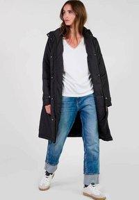 Zwarte lange pufferjas met een capuchon, voorzien van een rits- en drukknoopsluiting, gedragen over een witte V-hals T-shirt en lichtblauwe opgerolde spijkerbroek.