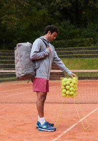 Mochila cinza, casaco cinza claro, calções cor-de-rosa, sapatos azuis. Cesto amarelo cheio de bolas de ténis num campo de terra batida. Ambiente externo com vegetação.