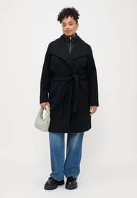Zwarte wollen mix coat met een brede kraag, een ceintuur in de taille en voorzakken, gedragen over een aansluitend zwart shirt en wijdvallende jeans.