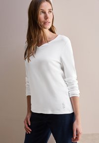 Witte katoenen top met lange mouwen, een bootnek en geborduurde details aan de zoom, gecombineerd met donkerblauwe broeken. Glad textuur.