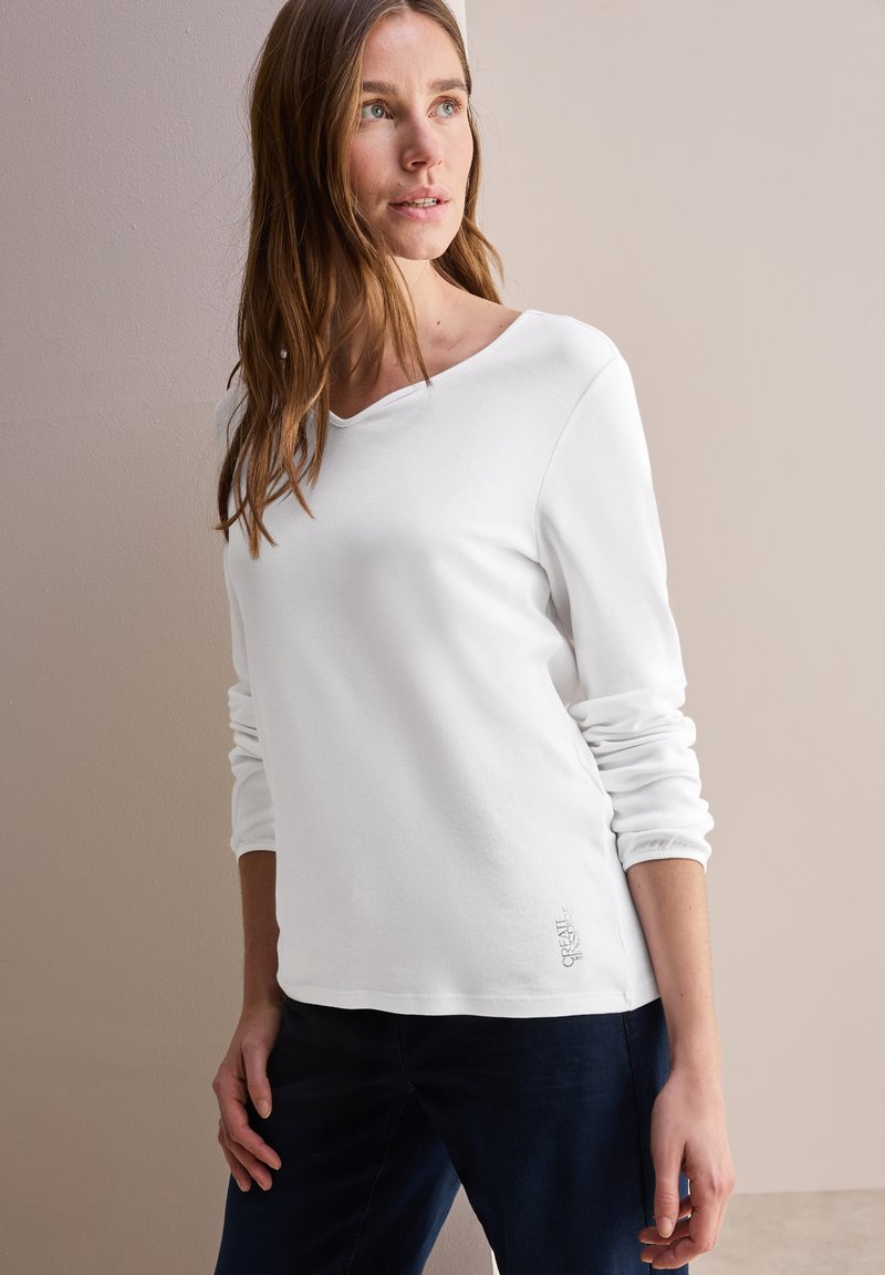 Witte katoenen top met lange mouwen, een bootnek en geborduurde details aan de zoom, gecombineerd met donkerblauwe broeken. Glad textuur.