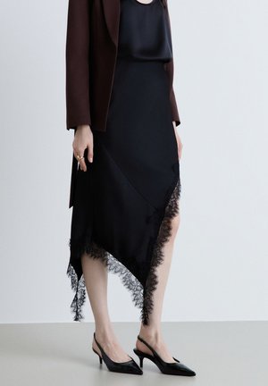 Donna che indossa una gonna asimmetrica nera con orlo in pizzo, top in raso nero, giacca marrone e tacchi slingback neri, in piedi su un pavimento semplice.